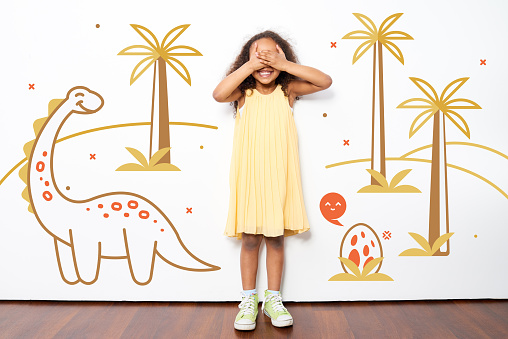 Positive excited African girl covering eyes with hands and happy about visit to Dinosaur park, funny dinosaur, egg and palms drawing behind her