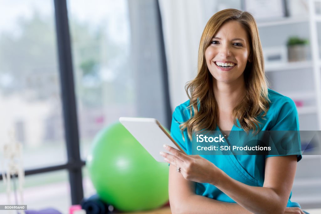 Portrait of beautiful physical therapist Beautiful mid adult Caucasian physical therapist holds a digital tablet. The tablet contains a patient's treatment plan. She is smiling cheerfully at the camera. Physical Therapist Stock Photo