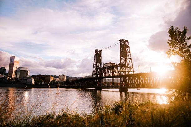 Portland's Steel Bridge, spanning the Willamette river and connecting the the waterfront Eastbank esplanade.  The sun sets behind the structure, casting warm light onto the cityscape.