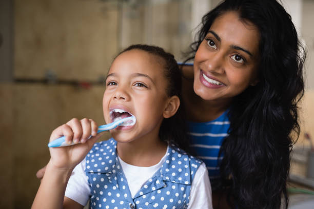 smiling mother standing by daughter brushing teeth in bathroom - human teeth child smiling family imagens e fotografias de stock