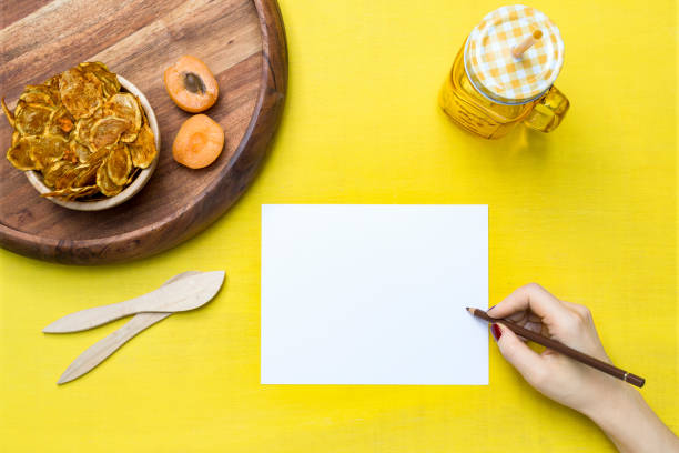 istock Yellow tabletop scene with female hand writing on a white paper with zucchini chips and apricots 800957576