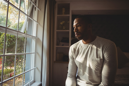 Thoughtful man sitting by window in bedroom at home
