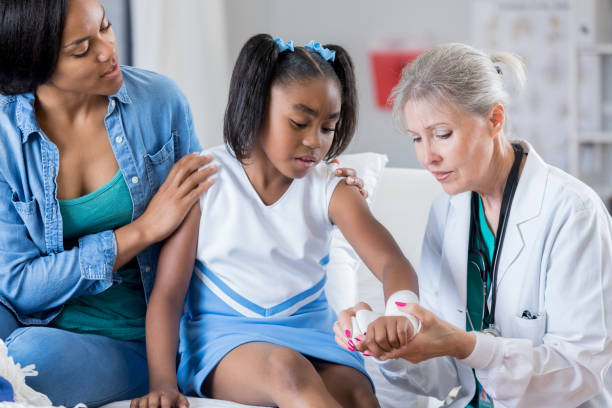 Doctor treats young injured cheerleader's wrist Senior Caucasian emergency room female doctor wraps young female African American cheerleader's injured wrist. The girl's mother is comforting her. emergency medicine stock pictures, royalty-free photos & images