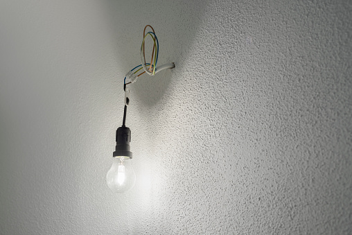 Illuminated halogen incandescent light bulb in a black plastic lamp holder socket with power cables protruding from a wall with white rough plaster