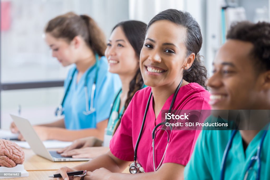 Medical student smiles at camera in class Attractive young African American medical student smiles at the camera.  She is sitting in the middle of three other students at a strings of desks. Two other students are looking forward and smiling and another is writing at her desk. Nurse Stock Photo