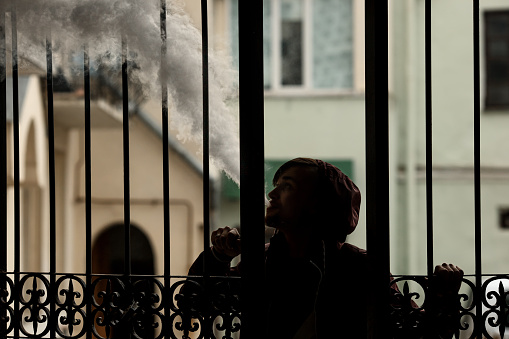 Vape man. Silhouette of a young white guy in the hood vaping and letting off steam from an electronic cigarette near a vintage metal gate. Lifestyle.