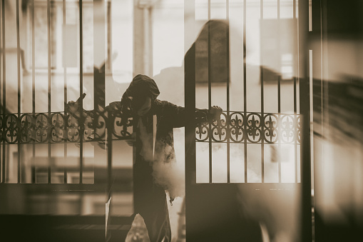 Vape man. Double exposure. Silhouette of a young white guy in the hood vaping and letting off steam from an electronic cigarette near a vintage metal gate. Lifestyle.