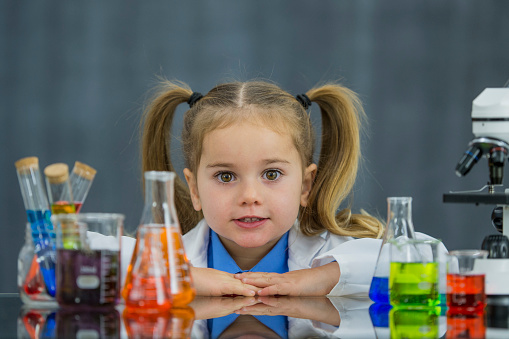 A female Caucasian toddler is dressed up as a scientist, wearing a lab coat and bow tie. She is standing behind a desk. The desk has various liquid-filled beakers and a microscope on it. She is looking forward.