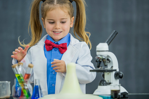 A female Caucasian toddler is dressed up as a scientist, wearing a lab coat and bow tie. She is standing behind a desk. The desk has various liquid-filled beakers and a microscope on it. She is watching a foamy reaction from one of the beakers of liquid.