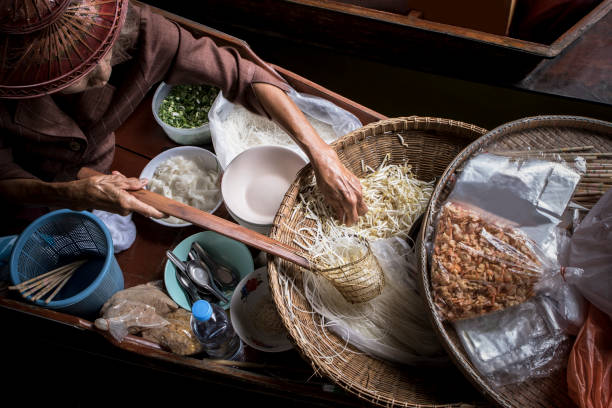 jeune femme de faire à manger aux nouilles thaïlandaises en naviguant dans le marché local de bateau flottant - thai cuisine photos et images de collection