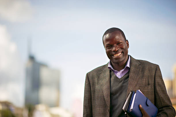Happy African Businessman Friendly Black African Businessman smiling holding files with a building and cloud sky backdrop Nairobi Kenya Africa kenyan man stock pictures, royalty-free photos & images