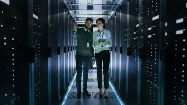 Photo of IT Engineer Shows Working Data Center / Server Room to Female Chief Engineer who Holds Tablet Computer.