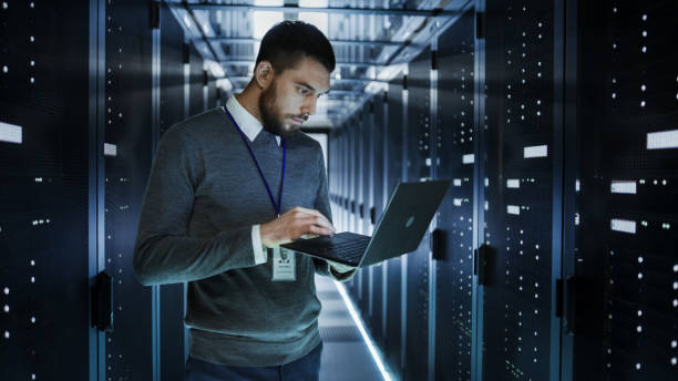 A man in a gray sweater and a ID badge stands in a server room, focusing on his laptop while surrounded by rows of dark server racks illuminated by LED lights.