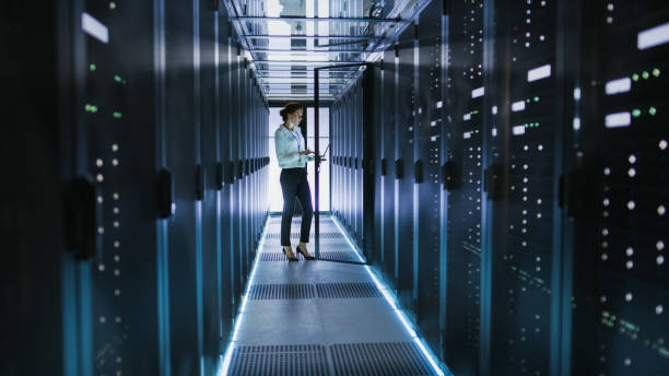女性サーバー技術者立つ行のラック マウント型サーバとデータ センターの廊下のキャビネットの横に。彼女は彼女のコンピューターに診断プログラムを実行 - women science service technology ストックフォトと画像