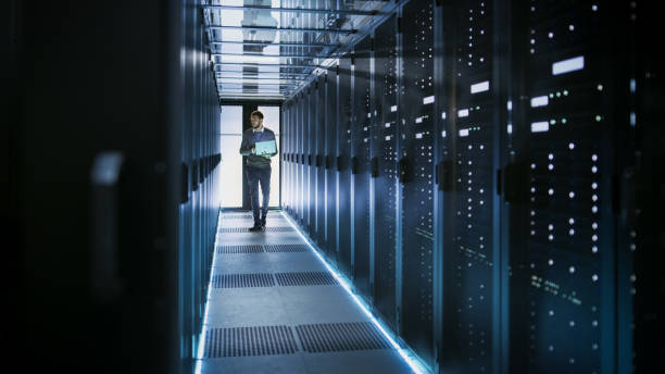 istock IT Technician Works on Laptop in Big Data Center full of Rack Servers. He Runs Diagnostics and Maintenance, Sets System Up. 802301324