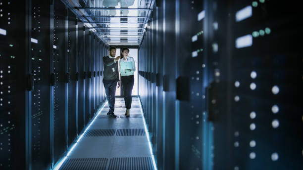 istock Female IT Technician and Male Server Engineer Talk and Discuss. They are in Working Data Center full of Rack Servers. Woman Holds Laptop. 802301334
