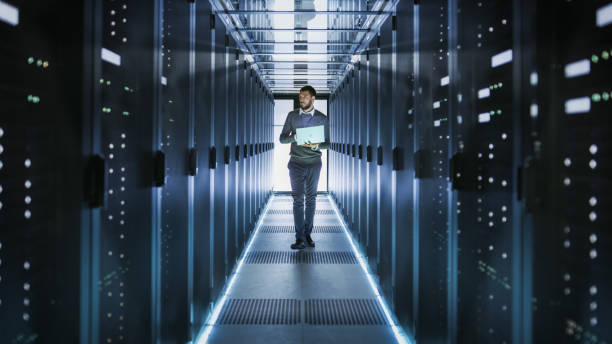istock IT Technician Works on Laptop in Big Data Center full of Rack Servers. He Runs Diagnostics and Maintenance, Sets System Up. 802301336