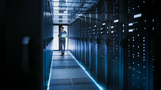 istock IT Technician Works on Laptop in Big Data Center full of Rack Servers. He Runs Diagnostics and Maintenance, Sets System Up. 802301342