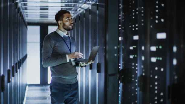 IT Technician Works on Laptop next to a Server Cabinet in Big Data Center. He Runs Diagnostics and Maintenance, Sets System Up. IT Technician Works on Laptop next to a Server Cabinet in Big Data Center. He Runs Diagnostics and Maintenance, Sets System Up. data center stock pictures, royalty-free photos & images