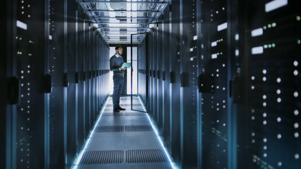 istock IT Technician Works on Laptop in Big Data Center full of Rack Servers. He Runs Diagnostics and Maintenance, Sets System Up. 802301366