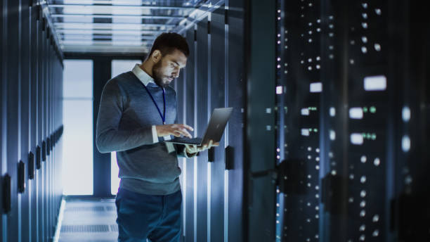 istock IT Technician Works on Laptop next to a Server Cabinet in Big Data Center. He Runs Diagnostics and Maintenance, Sets System Up. 802301372