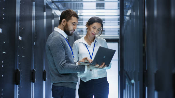 istock Male IT Specialist Holds Laptop and Discusses Work with Female Server Technician. They're Standing in Data Center, Rack Server Cabinet is Open. 802301374