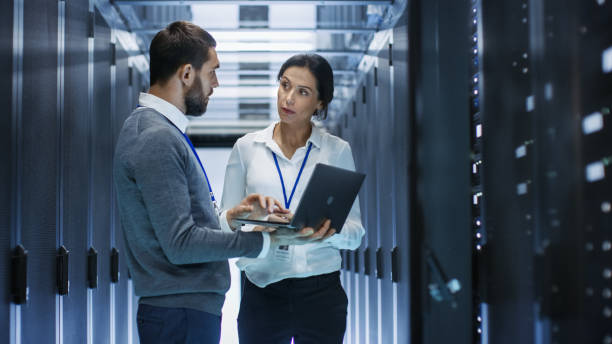 male it specialist holds laptop and discusses work with female server technician. they're standing in data center, rack server cabinet is open. - network server rack computer mainframe imagens e fotografias de stock