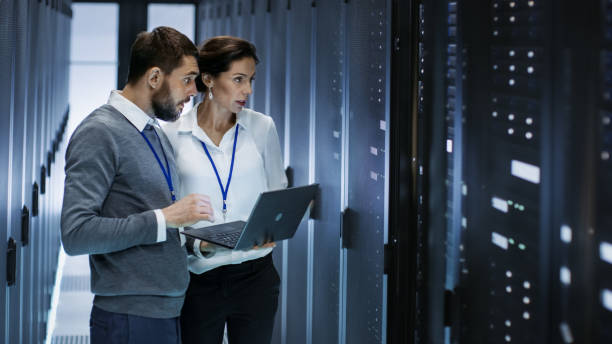 istock Male IT Specialist Holds Laptop and Discusses Work with Female Server Technician. They're Standing in Data Center, Rack Server Cabinet is Open. 802301400