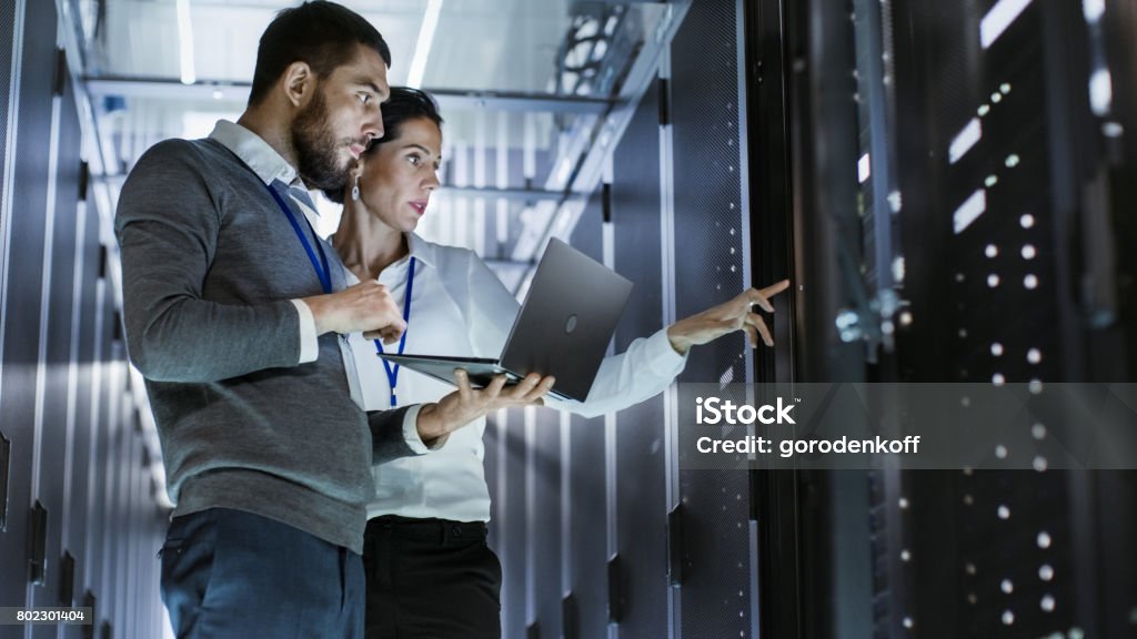 Male IT Specialist Holds Laptop and Discusses Work with Female Server Technician. They're Standing in Data Center, Rack Server Cabinet is Open. Technology Stock Photo