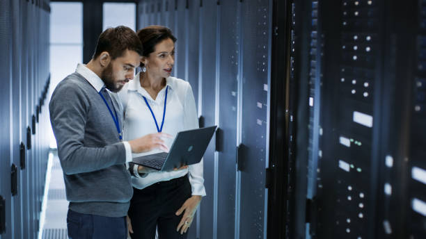 istock Male IT Specialist Holds Laptop and Discusses Work with Female Server Technician. They're Standing in Data Center, Rack Server Cabinet is Open. 802301406