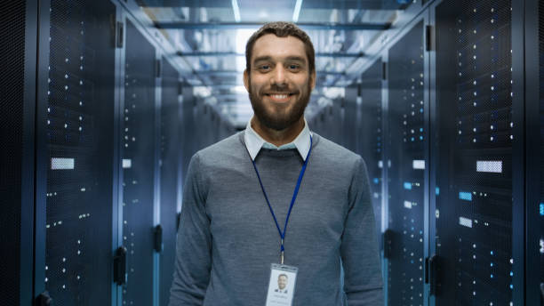 retrato de una curios, positivo y sonriente ingeniero de pie en medio de una sala de servidores del centro de datos grande. - profesional de informática fotografías e imágenes de stock