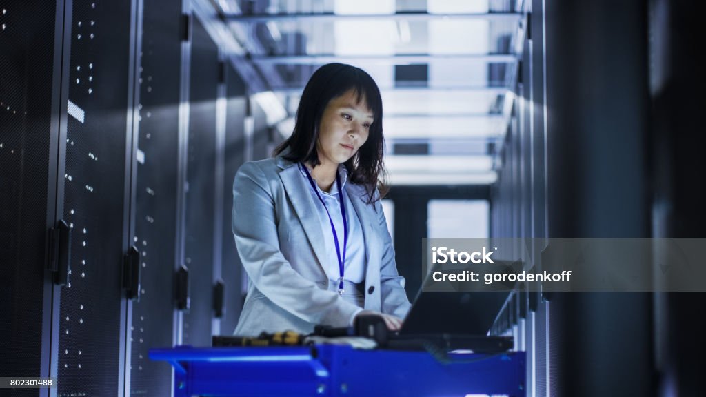 Asian Female IT Engineer Working on a Laptop on Tool Cart, She Scans Hard Drives.  She's in a Big Data Center Full of Rack Servers. Technology Stock Photo