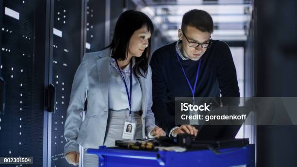 Caucasian Male And Asian Female It Technicians Working With Computer Crash Cart In Big Data Center Full Of Rack Servers Stock Photo - Download Image Now