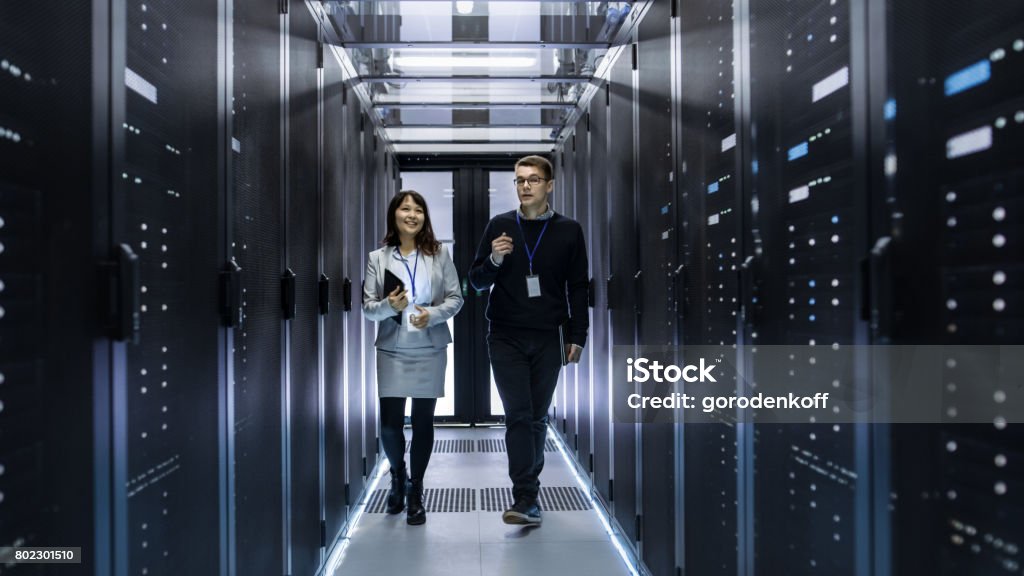 Caucasian Male and Asian Female IT Technicians Walking through Corridor of Data Center with Rows of Rack Servers. They Have Discussion, She Holds Tablet Computer. Data Center Stock Photo
