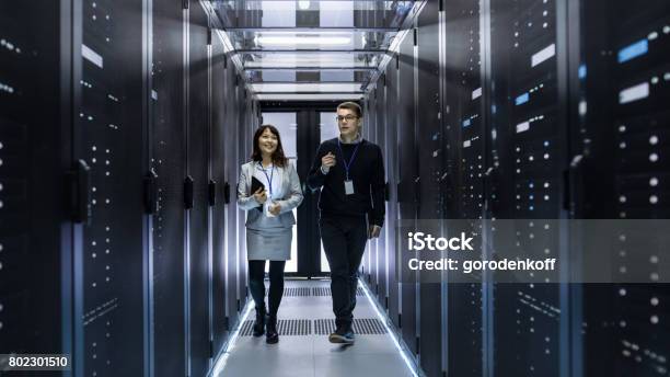 Caucasian Male And Asian Female It Technicians Walking Through Corridor Of Data Center With Rows Of Rack Servers They Have Discussion She Holds Tablet Computer Stock Photo - Download Image Now