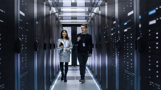 кавказские мужчины и азиатские женщины-техники,покатясь по коридору центра обработки данных с рядами серверов стойки. у них есть обсуждени - network server data center installing стоковые фото и изображения