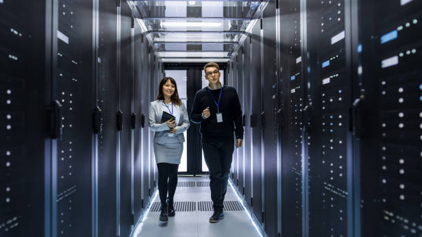 caucasienne hommes et asiatiques femmes techniciens marchant dans le couloir du centre de données avec des rangées de serveurs en rack. ils ont discussion, elle est titulaire d’ordinateur tablette. - network server data center installing photos et images de collection