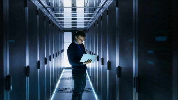 istock Male IT Engineer Works on a Laptop in front of Server Cabinet at a Big Data Center. Rows of Rack Servers are Seen. 802302950
