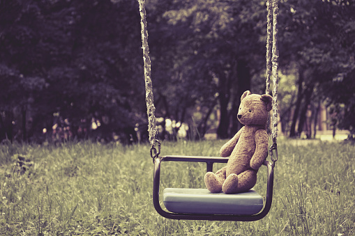 Old toy teddy bear sitting on swing in park, retro style, toned effect