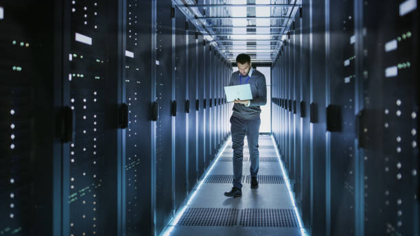 istock IT Technician Works on Laptop in Big Data Center full of Rack Servers. He Runs Diagnostics and Maintenance, Sets System Up. 802317154