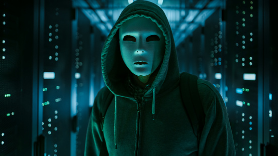 Masked Hacker in a Hoodie Standing in Corporate Data Center with Rows of Working Rack Servers.