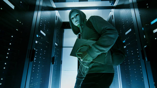 istock Low Angle Shot of a Masked Hacker in a Hoodie Sneaking Through Corporate Data Center with Rows of Rack Servers. 802320774