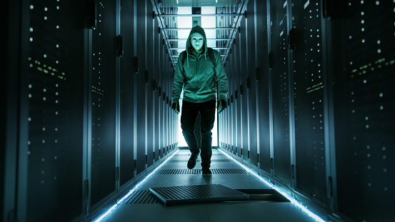 Hacker con capucha en una máscara camina a través de trabajo centro de datos con piso Abra la escotilla en medio de ella. photo
