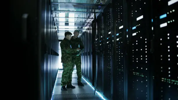 Photo of In Data Center Two Military Men Work with Open Server Rack Cabinet. One Holds Military Edition Laptop.