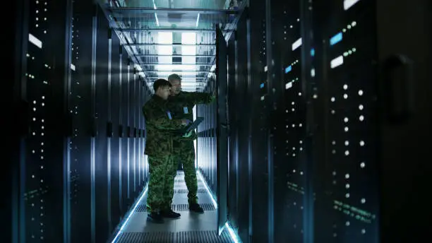 Photo of In Data Center Two Military Men Work with Open Server Rack Cabinet. One Holds Military Edition Laptop.