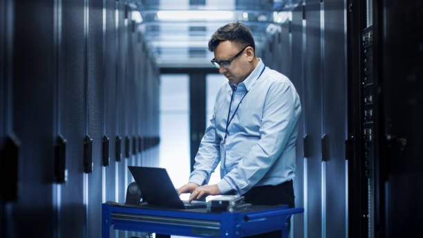 istock In Data Center IT Engineer Works on Crash Cart Laptop. We See Rows of Server Racks and One Cabinet is Open. 802320924