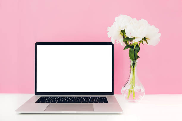 istock Laptop with white blank screen and flowers in vase on table on pink background. mock up 802321364