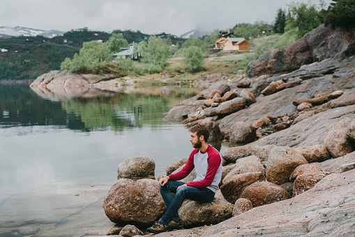 Young Caucasian man  sitting on rock and looking at scenic fjord in Norway