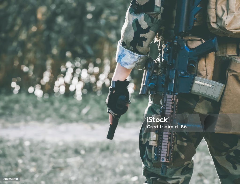 The soldier in the performance of tasks in camouflage and protective gloves holding a gun. The soldier in the performance of tasks in camouflage and protective gloves holding a gun. War Zone. Airsoft - Sport Stock Photo