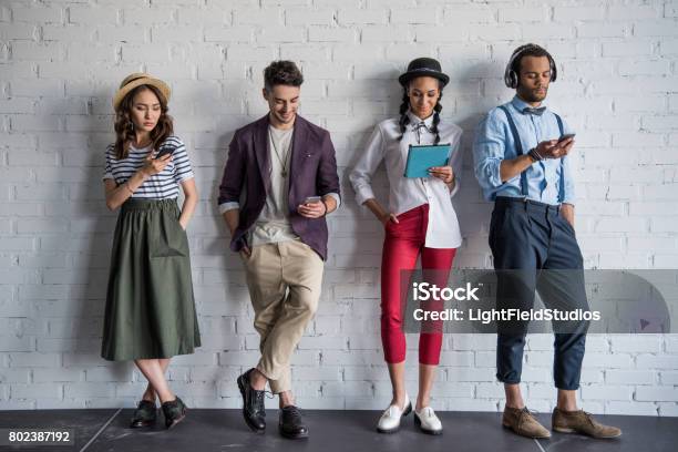 Young Stylish Friends Using Digital Devices While Standing Near Brick Wall Stock Photo - Download Image Now